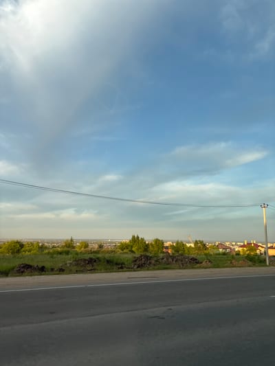The image depicts a landscape view of a suburban area with a road in the foreground and a city in the background, under a partially cloudy sky. The road is asphalt with a white line marking its edge. Adjacent to the road is a grassy slope with some piles of dirt. Beyond the slope, a line of trees separates the foreground from a vista of buildings with red and yellow roofs, characteristic of residential areas. A construction crane is visible in the distance, suggesting ongoing development. The sky is a mix of blue and white clouds, with some areas appearing bright, possibly indicating sunlight. A power line stretches horizontally across the mid-ground. The scene is set in the city of Plodopitomnik, Russia, and appears to be captured during daylight hours, likely late afternoon or early evening given the lighting. There are no people or animals visible in the frame.