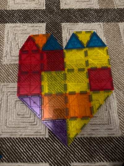 The scene, situated in Taastrup, Denmark, displays a heart-shaped construction formed from translucent magnetic building tiles. This object rests on a textured, light-colored woven textile surface, consistent with a rug or carpet, which features a repeating geometric pattern of intersecting dark and light stripes.
The heart comprises multiple individual tiles. The upper left lobe is predominantly formed by red square tiles and two red triangular tiles at its apex, with an additional orange triangular tile positioned at the very top center. The upper right lobe is primarily composed of yellow square tiles and two blue triangular tiles at its apex. The central and lower portions of the heart incorporate a mix of yellow, orange, red, and purple square and triangular tiles. Specifically, the lower left point of the heart is defined by a purple triangular tile, and the lower right point by another purple triangular tile. Each translucent tile exhibits internal grid patterns and small, dark-colored magnetic connectors along its perimeter. No people, discernible actions, or interactions are present in the image. The lighting is even and ambient, without casting distinct shadows.