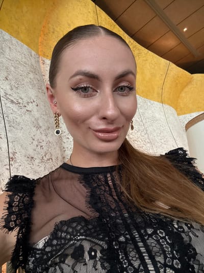 This is a close-up, vertical, selfie-style photograph of a woman with light skin and blonde hair. She is wearing a black lace top with a sheer overlay on the neckline and chest. Her hair is long and straight, falling over her shoulder. She has on makeup, including eyeshadow, eyeliner, and lipstick. She is also wearing earrings. One earring is a gold chain with a dark circular pendant. She has two ear piercings on her left ear, one with a small stud and the other with the dangling earring.

The background is abstract and textured, with areas of yellow and white. The yellow area has a ribbed texture, suggesting it could be part of a decorative architectural element or a piece of furniture. The white area appears to be a textured surface, possibly stone or plaster. The lighting suggests an indoor setting, possibly with artificial lights. No specific time of day or weather conditions are discernible. No visible text is present in the image. The woman's expression is neutral, with her gaze directed towards the camera.