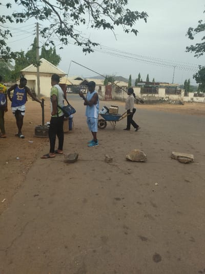 The image shows a scene on a dirt road in New Karu, Nigeria, located at the coordinates 8.993 latitude and 7.57 longitude. Four adults are standing or walking on the road. There are three men and one woman. One man is wearing a blue Lakers jersey, one is wearing a white T-shirt, one is wearing a blue tank top, and the woman is wearing a blue and white blouse. The woman is pushing a wheelbarrow containing items. The men are looking in different directions, and the woman is looking ahead. There are some large rocks scattered on the road. The environment is dusty and dry, with some buildings visible in the distance. The day is cloudy. The image was likely taken with a smartphone.