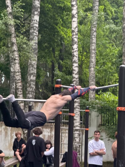 A man is performing a calisthenics routine on a horizontal bar outdoors, with other individuals standing and sitting nearby. The man is shirtless, wearing black pants and grey socks, and is mid-movement, with his body extended horizontally. His arms are wrapped in red and black patterned sleeves. The scene is set in a park-like area with birch trees in the background and a fence. Several young people are visible in the lower portion of the image, some sitting on a bench and others standing. One man in a white t-shirt stands with his arms crossed, facing the camera. This location is identified as Dmitrov, Russia, likely a public outdoor fitness area or park.