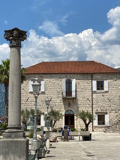 A stone building with a red tile roof sits under a partly cloudy blue sky in Perast, Montenegro. A large, decorative stone column stands in the foreground to the left, with a palm tree behind it. In front of the building, there is a paved courtyard with several objects including street lamps, benches, planters with small trees, and busts on pedestals. A set of ornate stairs leads up the right side of the building. The building has multiple windows with white shutters and a balcony above the main entrance. The overall scene appears to be a sunny day in a historic town.