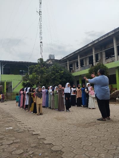 A group of approximately 25 young girls, all wearing headscarves and long dresses, stand in two lines facing a woman wearing a blue shirt and black pants in front of a partially-constructed building. The woman is gesturing with her left hand while looking at the girls. The scene is outdoors in Cangkuang, Indonesia, located at the coordinates -7.065 latitude and 107.551 longitude. There is a tall, white tower in the background with wires connected to it, a large, unfinished building with visible structural framing, and a green and brown leafy tree in the center of the image. The ground is covered with a pattern of gray, brick-like pavers. The sky is overcast with a light gray color.