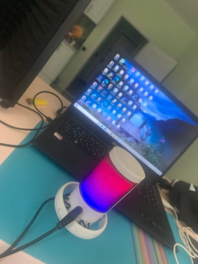 The image shows a close-up, angled shot of a laptop and a small, cylindrical speaker with colorful LED lights. The laptop is open, with its screen displaying a desktop background featuring a landscape with a mountainous region and a body of water. The keyboard is visible, and the laptop appears to be black.
The speaker is white at the top and bottom, with a mesh section in the middle that is illuminated with a gradient of pink and blue light. It is connected by a black wire to an unseen source. The speaker sits on a white base with a circular cutout.
The scene is set on a bright blue surface, possibly a desk or table, with a thin, striped mat underneath. In the background, a blurred interior is visible, suggesting a room. There are hints of furniture and possibly a kitchen area.
The overall impression is one of a personal workspace or entertainment setup. The lighting suggests it could be daytime, but the indoor setting makes it difficult to ascertain the time of day or weather. There are no people visible in the image. The focus is on the electronic devices. The image appears to be taken from a low angle, looking upwards at the subjects. No text is clearly legible in the image.