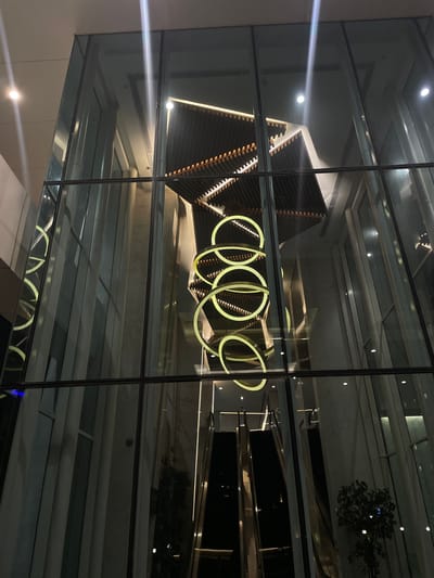 The image captures the interior of a modern building, likely a hotel lobby or a commercial space, viewed from the exterior through a large glass facade. The scene is dimly lit, with the primary light sources being elegant, circular pendant lights that hang in a cascading arrangement in the center of the atrium. These lights are a soft yellow hue and create a striking visual focal point against the darker backdrop.
The architecture features clean lines and geometric shapes. Above the pendant lights, a multi-tiered ceiling structure with wooden slats and integrated lighting is visible, adding a sense of depth and sophistication. The walls appear to be clad in a light-colored material, possibly marble or a similar stone, which reflects the ambient light.
In the lower portion of the image, a set of escalators is visible, suggesting the building's functionality as a multi-level space. The escalators are dark in color, with metallic trim, and appear to be in operation or ready for use.
Reflections on the glass facade create a layered effect, showing the interior elements superimposed on hints of the exterior environment, though the exterior is largely indistinct due to the low light conditions. The overall impression is one of contemporary luxury and a well-designed, atmospheric interior space. The building's facade is divided into a grid of large glass panes framed by dark metal mullions.