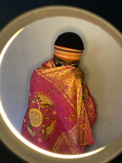 A person is positioned centrally within the illumination of a ring light, set against an unadorned, light-toned background. The individual's face is partially obscured by a black bandana displaying white patterns, covering from the nose down. Above the bandana, a striped headband, featuring horizontal bands of orange, brown, and a light contrasting color, is worn across the forehead. Dark, tightly curled hair is visible atop the head. The subject is draped in a vibrant pink fabric, rich with intricate gold patterns and embroidery. Prominent embellishments on the visible left side of the garment include clusters of white pearls, sequins, and gold beads arranged in ornate designs. A gold-colored trim with a geometric pattern borders one edge of the draped fabric. One hand is visible, holding the fabric up near the face, further concealing the lower facial features. A ring is present on a finger of this hand. The subject is directly facing the viewer. No visual elements within the image content confirm the requested location of Ngleshi Amanfro, Ghana.