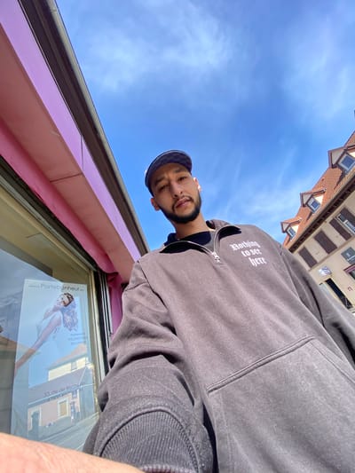 A low-angle photograph shows a person in Bischheim, France, looking down at the camera. The individual is wearing a dark-colored hoodie with the words "Nothing to see here" written in white text. They also wear a dark cap with a patterned brim. The backdrop features a clear, blue sky. In the lower part of the frame, a pink-colored building facade is visible, with a window displaying a poster. The poster features a woman and the website address "www.Portebonheur.eu," along with text indicating the location as "33, rte de Bischwiller 67800 BISCHHEIM" in Bischheim, France. Reflected in the glass of the poster is the image of a building. Tiled roofs and building exteriors are also visible in the upper section of the image.