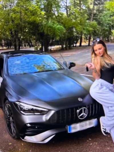 A young woman with blonde hair is leaning against the hood of a dark gray Mercedes-Benz vehicle. She is wearing a black sleeveless top and light-colored sweatpants. In her right hand, she is holding a car key. The car's front grille and headlight are visible, as is the Mercedes-Benz logo. The vehicle is parked on a paved surface, possibly a road or parking lot, with trees and foliage in the background. The image appears to have been taken outdoors during daylight.