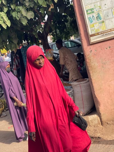 The image depicts a bustling outdoor market scene in Samaru, Nigeria, illuminated by direct sunlight. In the foreground, a woman wearing a vibrant red full-body abaya/hijab is positioned centrally, her face visible and looking towards the right of the frame. Her right hand holds a small black pouch or cap featuring a white Nike logo. Faint dark markings, consistent with henna or tattoos, are visible on her exposed right forearm. To her left, slightly behind her, another woman in a purple full-body abaya/hijab and a black face mask holds a bundle of light-colored sticks in her right hand.
On the far left, a pile of green and yellow bananas rests on the ground. Above, a large tree with dense green foliage provides shade, and various fabrics or garments are displayed hanging from a stand beneath its branches, alongside small packaged items suspended by strings. In the mid-ground, a male figure dressed in a light beige caftan is partially obscured, appearing to attend to several large white sacks on the ground, which are filled with light-colored grains or nuts. These sacks are situated alongside a textured, faded reddish-pink wall that forms the right boundary of the image. Affixed to this wall is a white rectangular sign displaying various cosmetic products and text including "TEL: 08107222094, UR BEAUTY". The background reveals parked vehicles on a street and additional blurred individuals, indicating a crowded urban environment.
