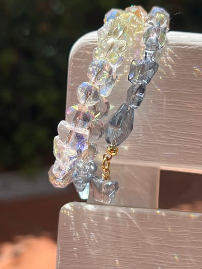 A close-up, eye-level shot focuses on two strands of iridescent and translucent blue glass beads, strung together with a gold clasp. The beads vary in shape and size, with some appearing faceted and others smooth and rounded. They are draped over a textured, white, possibly wooden surface that reflects light, creating rainbow-like refractions and glare across the image. The background is blurred, showing dark green foliage and a hint of reddish-brown. The scene is lit with strong, directional light, suggesting it is outdoors or near a window. The image does not contain information about the city of Bijela, Montenegro.