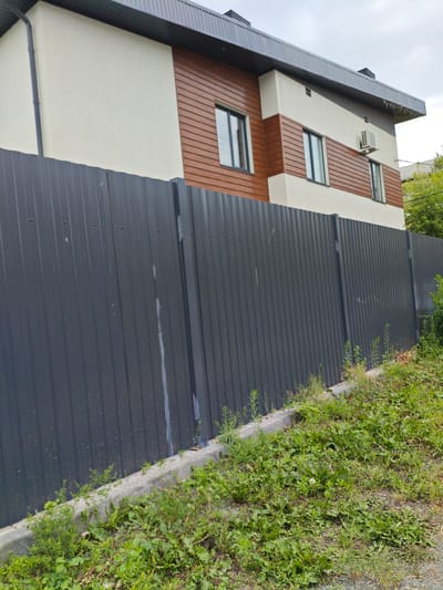 A medium-angle outdoor shot depicts a residential building and a dark gray corrugated metal fence. The building, with two visible stories, features a light-colored facade and horizontal brown paneling. Three rectangular windows are discernible, two on the upper story and one on the lower. An air conditioning unit is mounted on the building's exterior. The fence runs horizontally across the frame, partially obscuring the building's lower half. Its surface is textured by vertical ridges, and some faint white streaks are present. To the right of the fence, lush green vegetation, including grass and weeds, covers the ground. A concrete curb is visible along the base of the fence, separating it from the overgrown area. The sky above is overcast. The scene appears to be in an urban or suburban setting, possibly Chelyabinsk, Russia, given the architectural style. There are no people or animals visible in the image.