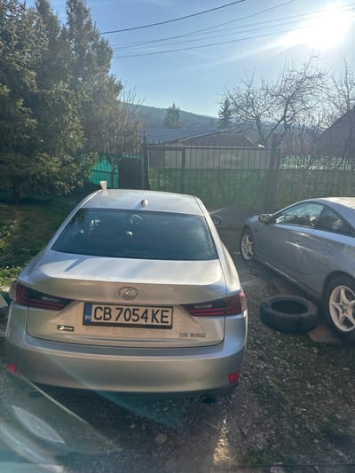 The image displays an outdoor scene in Katina, Bulgaria, under bright daylight conditions. Two vehicles are parked on a rough ground surface composed of gravel, dirt, and sparse grass. In the foreground, a silver Lexus IS 350 sedan is shown from its rear, angled slightly to the right. It bears a Bulgarian license plate, "CB 7054 KE," and an "F SPORT" badge on its trunk. A white paper cup is positioned on the car's roof, near the rear window and to the left of the shark fin antenna. To the right of the Lexus, a silver Toyota Celica coupe is parked, appearing to be in a state of maintenance or repair. Its front right wheel is missing, with a black tire (without a rim) lying on the ground directly adjacent to the vehicle, which is propped on wooden planks where the wheel should be. The visible wheels on the Celica are white, multi-spoke rims.

The background on the left features a line of tall, dark green coniferous trees. In the mid-ground, a dark metal fence, partially covered with green privacy screening, separates the vehicle area from another property. Beyond the fence, a building with a reddish-brown roof and what appear to be solar panels is visible. Further in the distance, a natural landscape of hills covered with a mix of evergreen and bare-branched deciduous trees extends under a clear blue sky. Overhead, thin power lines traverse the upper portion of the frame. The sun, prominent in the upper right quadrant of the sky, casts strong glare and shadows across the scene. No individuals are visible.