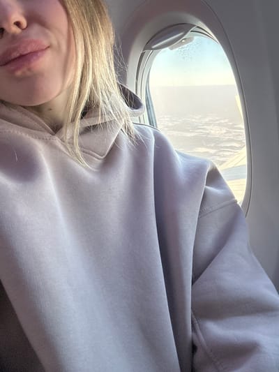 A blonde female is depicted in a close-up, seated inside an airplane cabin during daytime flight. Her lower face, including her mouth and chin, and light blonde hair are visible on the left side of the frame. She wears a plain, light lavender-colored hooded sweatshirt. To her right, an oval airplane window reveals an aerial view of an extensive layer of white clouds below, beneath a clear light blue sky. A portion of the airplane's wing, light in color, is visible at the lower right edge of the window. The window frame is a light gray with a small operating latch at the top. The scene depicts an in-flight journey, contextually associated with Teigebyen, Norway.