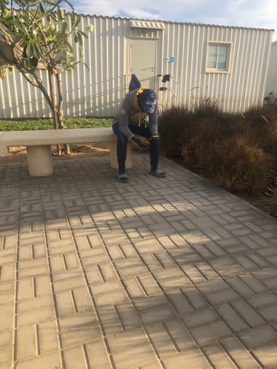 An outdoor scene in النعيلة, Saudi Arabia, depicts a single individual seated on a light-colored stone bench. The person, wearing a grey hooded sweatshirt with yellow accents, dark blue jeans, and light grey slide sandals, is looking down at a dark-colored smartphone held in both hands. White earbuds are visible, connected to the device. The bench is situated on a paved surface of light-colored rectangular pavers laid in a repeating pattern. To the left of the bench, a small tree with green leaves stands next to a patch of green grass. To the right of the paved area, a section of brown ornamental grasses is visible. In the background, white, corrugated metal buildings feature a white door and a rectangular window. A dark garment and other clothes are hanging on a white rack or railing next to the building door. The scene is illuminated by natural light, casting distinct shadows on the paved surface, under a partly cloudy sky.