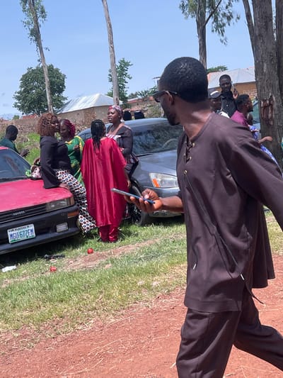 The image depicts an outdoor scene in Wumat, Nigeria, where a group of people are gathered near several vehicles. In the foreground, a man wearing a dark brown, loose-fitting garment walks away from the camera, holding a smartphone. Behind him, a dark sedan is visible. To the left, a red Toyota car is parked, with a license plate reading "PLATEAU MGU-672FM". A woman in a black top and patterned pants is leaning on the hood of the red car, interacting with other individuals. A woman in a bright red garment stands near the red car, facing away from the camera. Further back, other people are seen standing around, with buildings and trees forming the backdrop under a clear sky. The ground is a mix of grass and dirt.