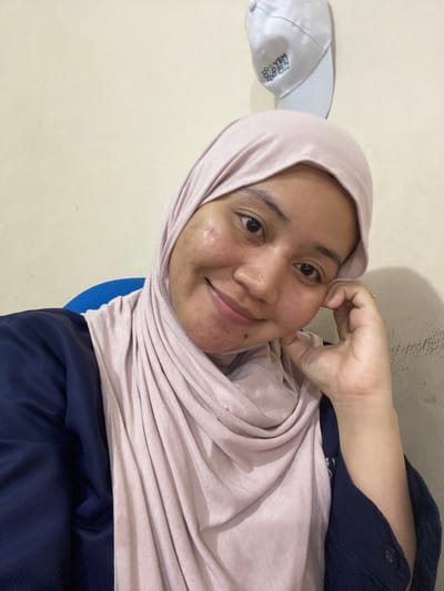 A young Indonesian woman, appearing to be in her early twenties, is captured in a close-up, selfie-style shot. She is wearing a light pink hijab and a dark blue top. Her right hand is resting on her cheek, and she has a gentle smile on her face, conveying a relaxed and content emotion.
The background is a plain, light-colored wall, with a white cap visible on the left side of the image. The cap has some text and a logo printed on it. The lighting suggests an indoor setting, possibly during the daytime. The overall scene is casual and intimate, typical of a personal photograph.
The visible text on the cap reads "YBM" and "BOYPEN". There are no obvious cues about the time of day or weather conditions beyond what is implied by the indoor setting. The location context provided is Banjarbaru, Indonesia.