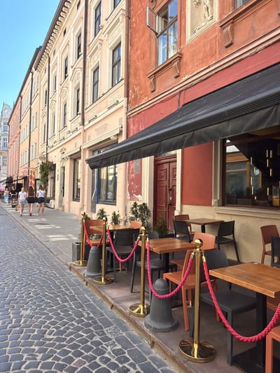 A low-angle, vertically oriented, full shot captures an outdoor cafe scene along a cobblestone street in Lviv, Ukraine. The buildings lining the street feature ornate, pastel-colored facades with many windows and decorative architectural elements. An awning extends from one of the buildings, providing shade over a seating area with tables and chairs. Red ropes on stanchions guide patrons, suggesting a queue or a designated walkway. Several people are visible, walking along the street and sitting at the tables. The scene appears to be set during daytime, with clear blue skies visible in the background. The atmosphere is casual and relaxed, typical of a leisurely day in a European city. The cobblestone street indicates an older, historic urban area. No text is clearly legible in the image.