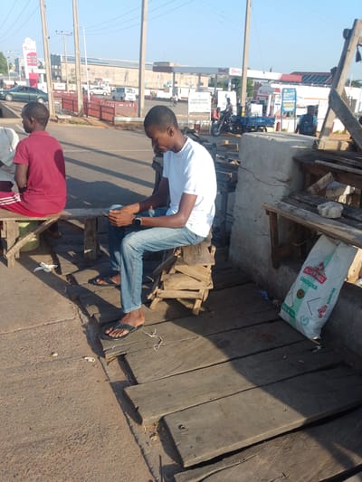 In Tumfure, Nigeria, two young men are seated outdoors in the daytime. The man on the left, wearing a red t-shirt and striped athletic pants, is sitting on a wooden bench. To his right, another man in a white t-shirt and blue jeans sits on a makeshift wooden stool. He is holding what appears to be a mobile phone. In the background, a gas station is visible with cars and a motorcycle. A bag of semolina is placed on the ground near the man in the white t-shirt. The scene is illuminated by natural sunlight, casting shadows on the ground.