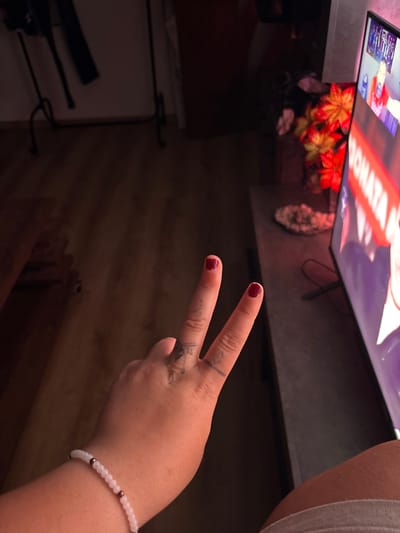 A hand with a beaded bracelet displaying the peace sign is in the foreground. Red nail polish is on the fingernails. Small tattoos are on the fingers. A TV is on a table in the background, displaying a floral arrangement in a vase. Clothes are hanging on a rack. The location is unknown.