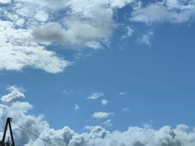 The image depicts a bright blue sky filled with scattered cumulus clouds. The clouds vary in size and density, with some appearing fluffy and white, while others are darker and more defined, suggesting depth and varying light conditions. In the lower left portion of the frame, a dark pole, likely a utility pole, is visible, with a wire extending diagonally across the lower section of the image. The pole has some foliage at its base. The overall scene is an outdoor environment under a daytime sky. The location is the city of Salacgrīva, Latvia.