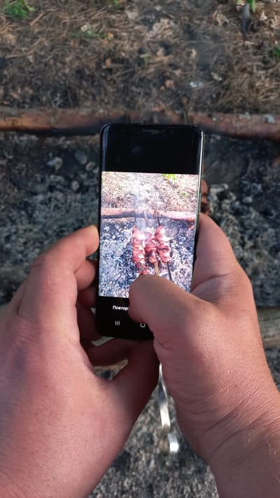 The image shows a close-up of a person's hands holding a smartphone. The smartphone screen displays a picture of meat skewers being cooked over coals. The background appears to be outdoors, with dry vegetation and what looks like a fire pit. There are no discernible landmarks or specific details to place the location within the city of Obukhiv, Ukraine.
