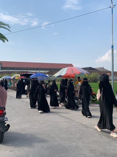 A group of women, mostly wearing black niqabs and abayas, walk down a paved road in Tanjungsepreh, Indonesia. Some are carrying bags. Several colorful umbrellas are set up, presumably for vendors. A man in a yellow shirt stands behind a cart. A motorcycle is parked on the left side of the road. In the background, there is a red-roofed building, lush green vegetation, and a clear blue sky with scattered clouds. A lamppost with blue and white accents stands to the right of the frame.