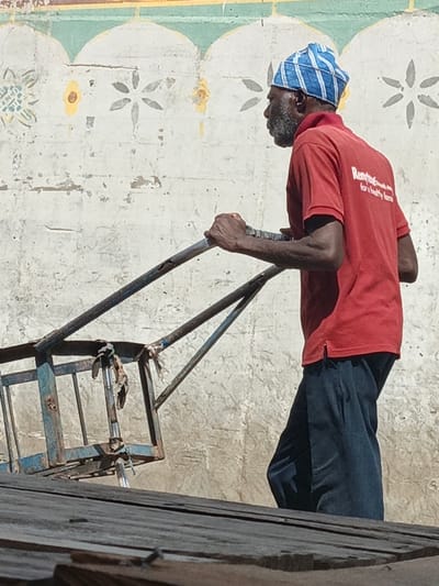 An adult male with dark skin and a graying beard, wearing a blue and white patterned skullcap, a red polo shirt with the text "Rentog... for a healthy home" on the back, and dark trousers, is depicted in Fagge C, Nigeria. He is actively pushing or pulling a worn, light-blue metal cart with visible rust and attached fabric scraps. His hands grip the cart's handlebars. The background consists of a weathered, light-colored wall adorned with a faded decorative pattern of arched segments and floral motifs in yellow and gray. Below the subject and cart, a dark, textured surface, likely wood or corrugated metal, occupies the foreground. The scene is brightly lit by natural sunlight.