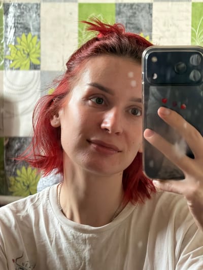 A woman with red hair is taking a selfie with a black smartphone in a clear case decorated with red hearts. She is wearing a white t-shirt and a silver chain necklace. The background is a table covered with a checkered tablecloth featuring green floral patterns. The location, Заболотово, Russia, is not discernible from the image.