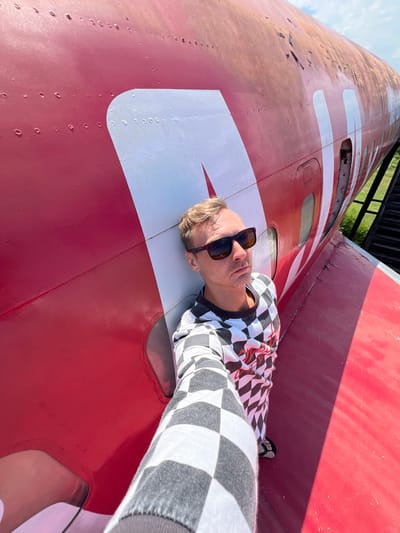 A man is lying on his back on the fuselage of a red and white airplane. He is wearing sunglasses and a black and white checkered long-sleeved shirt. The plane appears to be outdoors, with a blue sky and some green foliage visible in the background. The sun is shining, casting a shadow from the plane's structure onto the fuselage. The man is looking up at the sky, with a relaxed expression on his face. The airplane is large and cylindrical, with several windows visible along its length. The red and white paint scheme is bold and distinctive. The overall scene suggests a leisurely or adventurous moment, perhaps a tourist posing with a unique piece of aviation history. There is no visible text in the image.