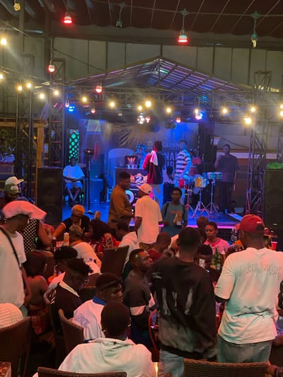 The image depicts a live music performance at an outdoor venue in Obosi, Nigeria. A stage with metal scaffolding is illuminated with blue and yellow stage lights. A band is performing on stage, including a drummer. A crowd of people, primarily African, are seated at tables, watching the performance. Some individuals are wearing casual clothing. The venue is also decorated with hanging string lights.