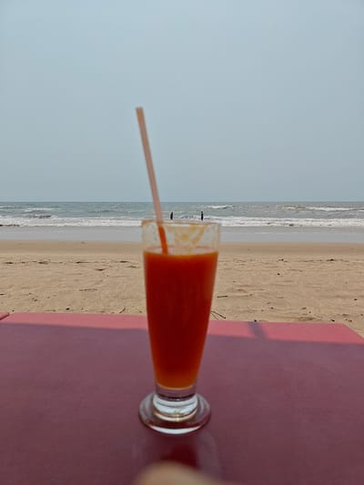 A glass of orange juice with a straw sits on a red table at Arambol beach, India. The beach stretches towards the horizon under a pale blue sky. Two people stand in the ocean, their figures small against the backdrop of waves. The sand is light brown and speckled with debris. The image is taken at eye-level.