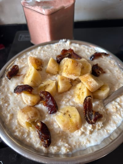A top-down close-up shot shows a stainless steel bowl containing oatmeal topped with sliced bananas and chopped dates. The oatmeal appears creamy, and the fruit is evenly distributed across the surface. A spoon rests partially submerged in the oatmeal. The background includes a blurred portion of a pink object and a black surface, possibly a stovetop. The location and people are not discernible from the provided image.