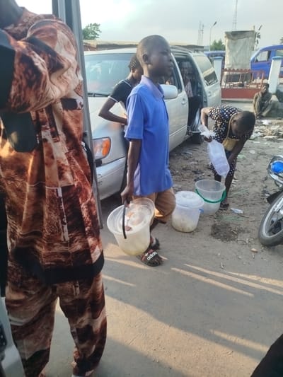 This is a street scene in Uduagba, Nigeria, with a silver minivan parked on the left. In the foreground, a person wearing a brown and white patterned garment is partially visible, facing away from the camera. To the right of this person, a young boy in a blue shirt and khaki pants is standing, holding a white plastic bucket with a black handle. The bucket appears to contain a light-colored liquid.

Behind the boy, a woman in a polka-dot dress is bent over, holding a clear plastic bag filled with ice cubes. In front of the woman are two more white plastic buckets, one with a green handle containing a similar light-colored liquid as the boy's bucket, and another smaller, lidded bucket with a white contents.

In the background, several other individuals are visible, including someone sitting on the ground on the right. Various objects, including trash and what appears to be a motorcycle, are scattered around the scene. The ground is dirt and gravel, indicative of a road or public area. The sky is overcast.