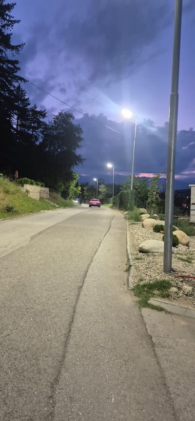 This image displays an outdoor street scene in Bania, Bulgaria, captured during twilight or dusk. The sky overhead is characterized by deep blue and purple hues with visible cloud formations, and a faint pinkish glow is discernible on the distant horizon, suggesting either sunset or ambient light.
The foreground features an asphalt road, exhibiting visible seams and patches indicative of construction or repair. The road appears to ascend or gently curve into the distance. To the immediate right, a concrete sidewalk is present, bordered by a curb. Adjacent to the sidewalk, a landscaped verge contains light-colored gravel, several large, rounded natural stones, and small green shrubbery. A tall, light-grey metallic pole, likely a streetlight standard, is prominently situated on the far right edge of the frame.
Multiple active streetlights illuminate the scene with bright white light, casting distinct lens flares, particularly from a large fixture in the upper right. Additional streetlights are visible further down the road.
On the left side of the road, a grassy embankment slopes upward, densely covered with dark trees and green bushes. Further up this slope, a low, light-colored retaining wall and a dark metal railing are visible.
Two vehicles are present on the road. A distinct red or magenta two-door sports car is centrally positioned in the middle distance, facing away from the camera. Further beyond this vehicle, two faint white headlights indicate another vehicle approaching.
The background reveals the faint, dark silhouette of mountains against the twilight sky, colored with subtle purple and pink tones. Dense vegetation lines both sides of the roadway, partially obscuring the distant landscape.