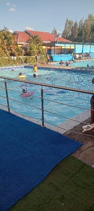 The image shows a swimming pool with a blue surface, surrounded by green grass and a blue mat on the edge. The pool is enclosed by a metal railing. There are several people in the pool, some are swimming and others are standing. A single person wearing black clothing is standing next to the railing, outside the pool, at the edge of the blue mat. They have one foot on the mat and one foot on the concrete. The pool is located at a resort in Athi River, Kenya. The weather appears sunny, with a single small cloud in the sky. The image is taken from the ground looking toward the pool. There are some buildings in the background. There are some trees around the pool. The image has been taken from a low angle. The pool is in the foreground of the image.