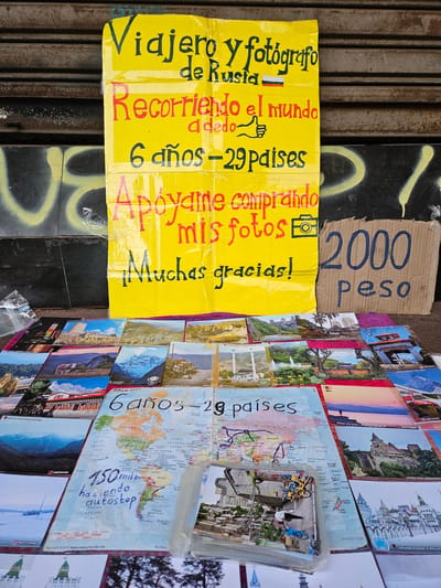 The image shows a street vendor's display in Osorno, Chile. A handwritten yellow sign advertises a Russian traveler and photographer who has traveled the world for 6 years, visiting 29 countries by hitchhiking ("a dedo"). The sign encourages support by buying their photos and offers thanks. Next to the sign is a cardboard sign with the price "2000 peso". Postcards, including images of various landmarks and world maps showing the traveled routes, are arranged on the side, potentially as samples of the photographer's work. The display sits against a backdrop of a metal roller door with graffiti and a dark surface.