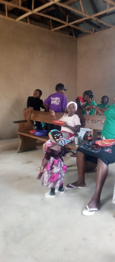 In an indoor setting, likely in Maigizo, Nigeria, a group of people are gathered, seated and standing. The room has unpainted concrete walls and a wooden-slatted ceiling. A long wooden bench runs along the back of the space, with several individuals seated on it.
In the foreground, a young child, dressed in a vibrant pink and purple patterned dress and a colorful headscarf adorned with lights, stands facing away from the camera. The child appears to be walking or dancing. Nearby, a young girl in a light-colored shirt and a white headscarf sits on the bench, holding a red bowl.
Other individuals are visible on the bench, including a person in a purple shirt with "Destiny Child" written on the back, and a person in a green shirt. A few people are engaged in conversation or observing the child. A table with plates, bowls, a metal cup, and a bottle of Coca-Cola is also present. The floor is made of concrete. The overall atmosphere seems to be casual and possibly part of a gathering or event.