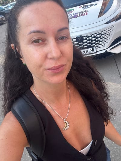 A selfie of a woman with dark curly hair and a silver necklace with a sun pendant is taken outdoors in Irkutsk, Russia. She is wearing a black vest over a white top. A white car with a "Suicide Squad" decal on the hood is visible in the background, with a license plate that reads "XP 38 RUS". The woman has on a black earbud in her left ear and a small green earring. The pavement and some trees are also visible in the background.