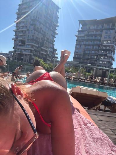 The image depicts a sunny day scene at a rooftop pool in Kâğıthane, Turkey, with two modern, high-rise apartment buildings prominently in the background under a clear blue sky. In the foreground, a person wearing a red bikini is lying on a pink towel on a sun lounger. Their back and buttocks are the main focus, with their legs crossed and slightly raised. To the left, another person, partially visible, is wearing glasses and a white cap. In the middle ground, several other individuals are seen near the pool, some sitting in the water and others on loungers. The pool area features wooden decking and numerous sun loungers, some occupied by people. The overall atmosphere suggests a relaxed, leisure setting.