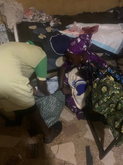 In Shagamu, Nigeria, a woman in a purple headscarf and patterned dress is tending to a newborn baby wrapped in a light-colored blanket. The baby is lying on a patterned green cloth, and a person wearing a yellow and green shirt is also present, leaning over the baby. The background shows a cluttered room with bedding, fabrics, and what appears to be medical supplies, including a disposable bed pad with some dark staining. The floor is tiled with a mosaic pattern.