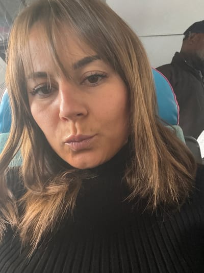 The image displays a close-up, front-facing portrait of an adult female, likely a selfie, taken indoors. The woman has medium brown hair with bangs styled across her forehead and falling to her shoulders. Her eyes are dark, and her lips are pursed in an expression resembling a "kissing" or "duck face." She is wearing a black, ribbed turtleneck sweater.
In the blurred background, to the left of the woman's head, a light blue seat headrest or cushion with a distinct pink border is visible. To the right, a dark-skinned male is partially visible, out of focus, wearing a dark jacket and a dark cap or hat. The environment suggests the interior of a vehicle, possibly a train, bus, or airplane, due to the visible seating and general cabin appearance.
The woman is directly engaged with the camera. There is no discernible interaction between the woman and the individual in the background. The specific location within the city of Lançon-Provence, France, cannot be determined from the visual information presented in the image.