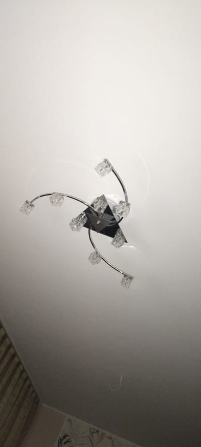 A modern, multi-arm chrome ceiling light fixture with crystal accents is mounted on a white ceiling. The fixture has a central black square base from which several curved chrome arms extend, each holding a clear, cube-shaped crystal. The overall impression is of a contemporary, decorative lighting element. The image appears to be taken indoors, possibly in a residential setting. No specific location in Aktobe, Kazakhstan can be determined from the visual information provided.