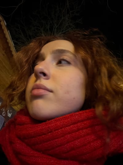 A woman with reddish-brown curly hair is seen from a low angle, wearing a thick red knit scarf. Her head is tilted slightly. She has fair skin, freckles, and her gaze is directed to the left of the frame. The background behind her head is dark, revealing the stark silhouette of bare tree branches. A dimly lit building is visible in the lower left corner.