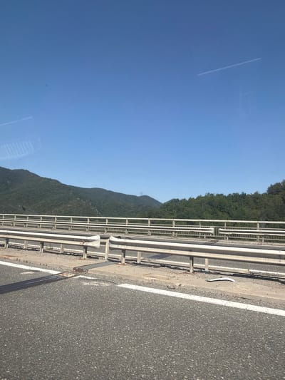 The image shows a view from a vehicle traveling on a highway, likely the Hemus highway near the town of Pravets, Bulgaria. The foreground consists of asphalt road with white lane markings. Beyond the road, a metallic guardrail system runs parallel to the direction of travel. The guardrail is composed of horizontal metal bars supported by vertical posts. In the background, a dense green forest covers rolling hills and mountains under a clear blue sky with a faint contrail from an aircraft. The lighting suggests it is daytime. No people or specific landmarks are visible to pinpoint the exact location beyond the general highway context in the region of Pravets, Bulgaria.