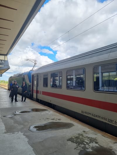 At a train station in Gorna Oryahovitsa, Bulgaria, a white and red train stands at a platform. A group of people in dark clothing are positioned next to the train. The platform is wet, with visible puddles. The sky is visible with blue patches amidst clouds. The station has a roofed waiting area, partially visible in the left of the frame.