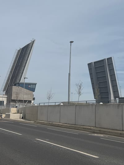 The image depicts a partially opened drawbridge located in Gorleston-on-Sea, United Kingdom. The bridge is constructed of gray concrete and metal, featuring two sections raised at angles to allow passage for marine traffic. A street, marked with white lines, runs alongside the bridge’s infrastructure. Several bare trees are visible along the bridge's length. A tall lamppost stands near one section of the bridge. A control tower-like structure is situated between the raised bridge sections. The sky is overcast.
