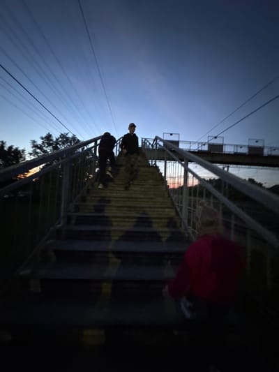 The image shows a low-angle view of a bridge, possibly a pedestrian overpass, in an urban or semi-urban area. Two people are walking on the bridge, their figures silhouetted against the evening sky. A third person, partially obscured, is situated closer to the camera on the bridge. The bridge's structure is metallic, with a textured surface that seems to have a star pattern. Overhead power lines are visible against the blue sky. Trees are present at the base of the bridge on the left. The location may be the city of Ферма, Russia, based on the instructions.
