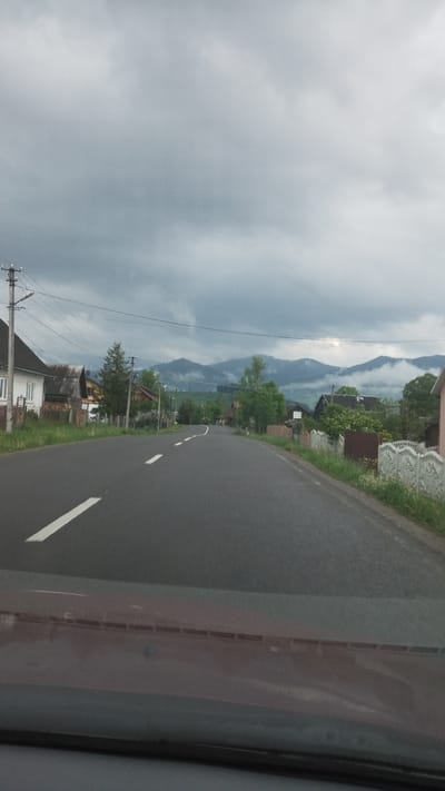 The image shows a view from inside a vehicle, possibly a car, traveling on a road. The road is paved and has lane markings. To the left, houses and buildings are visible, interspersed with trees and vegetation. In the background, mountains are present. The sky is overcast with heavy clouds. The scene is in the city of Lazeshchyna, Ukraine.