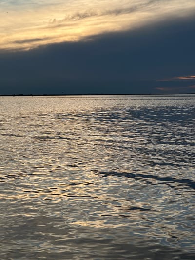 The image depicts a horizontal split scene, with the sky on the left and water on the right. The sky is a deep, dusky blue, transitioning to lighter hues with streaks of golden-yellow and white clouds, suggesting either sunrise or sunset. A faint, dark line along the horizon separates the sky from the water. The water is rippled, with sunlight glinting off the surface, creating a dynamic pattern of light and shadow. The ripples are elongated and create a textured, almost abstract effect on the water. The overall mood is serene and natural. There is no visible text in the image. The location context provided is Kokshetau, Kazakhstan, which could imply a large body of water such as a lake or a river within that region. The time of day is likely early morning or late evening due to the lighting conditions in the sky. The weather appears calm, with gentle ripples on the water.