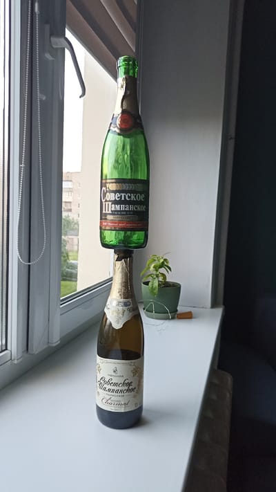 Two bottles of Soviet Champagne are stacked vertically on a windowsill. The top bottle is green, and the bottom bottle is a darker amber color. Both bottles have labels indicating "Soviet Champagne" and other text in Russian. A small potted plant with green leaves is to the right of the bottom bottle, with a strand of fairy lights wrapped around it. The windowsill is white and appears to be in an interior setting, with a window to the left showing an urban background with buildings and trees. The scene is well-lit, likely by natural light from the window.